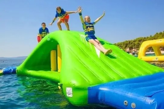Three people sliding on a green inflatable water slide in the ocean, wearing life vests.