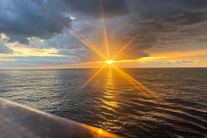 Sunset over a calm ocean with a cloudy sky and sun rays visible.