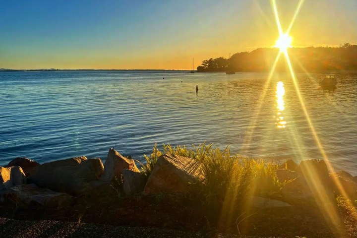 Sunset over calm lake with rocks in foreground and sun rays reflecting on water.