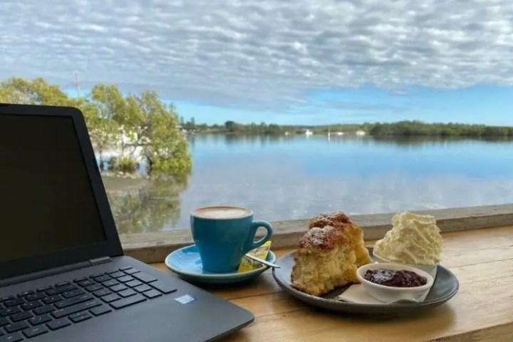 Laptop, coffee, scone, and cream on table by scenic lake view with cloudy sky.
