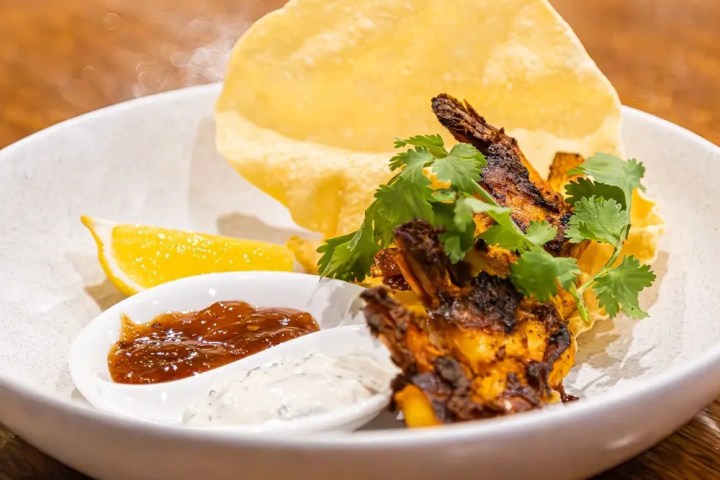 Grilled shrimp with herbs, lemon wedges, sauces, and a crispy tortilla on a white plate.