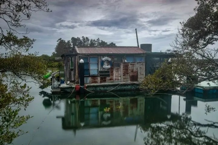 Rustic houseboat on a calm river with trees and overcast sky.