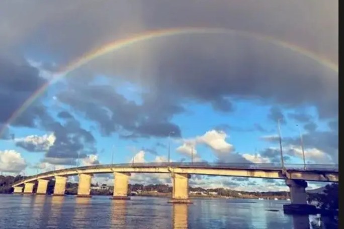 Rainbow over a bridge spanning a wide, calm river with cloudy sky.