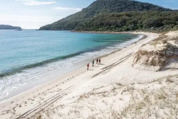 Beach with white sand, people walking, lush hills and blue sea.