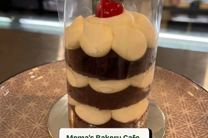 Layered chocolate cake in clear wrap with cream and cherry on top.